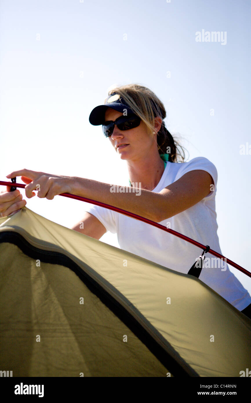 A blonde woman in sunglasses puts up a tent in the sun. Stock Photo