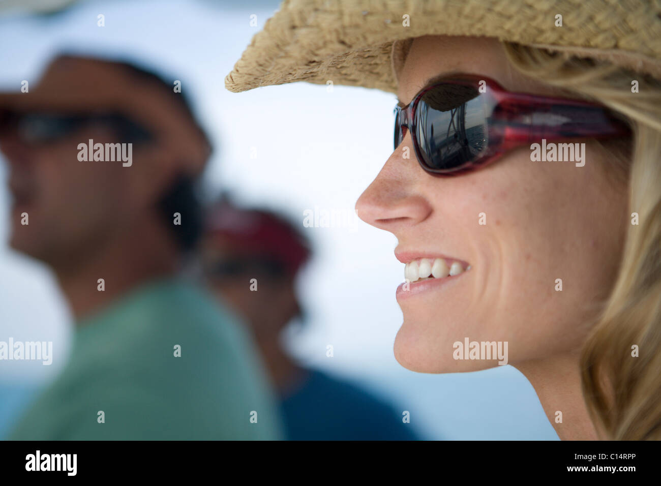 A close-up of a blonde woman as she looks out to the water wearing sunglasses and a straw hat. Stock Photo