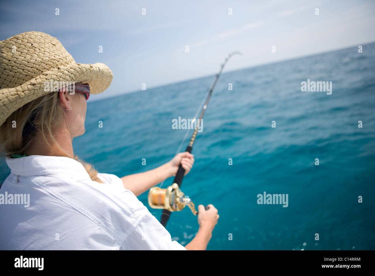 A blonde woman in a straw hat reels in a fish with blue wanter in the distance. Stock Photo