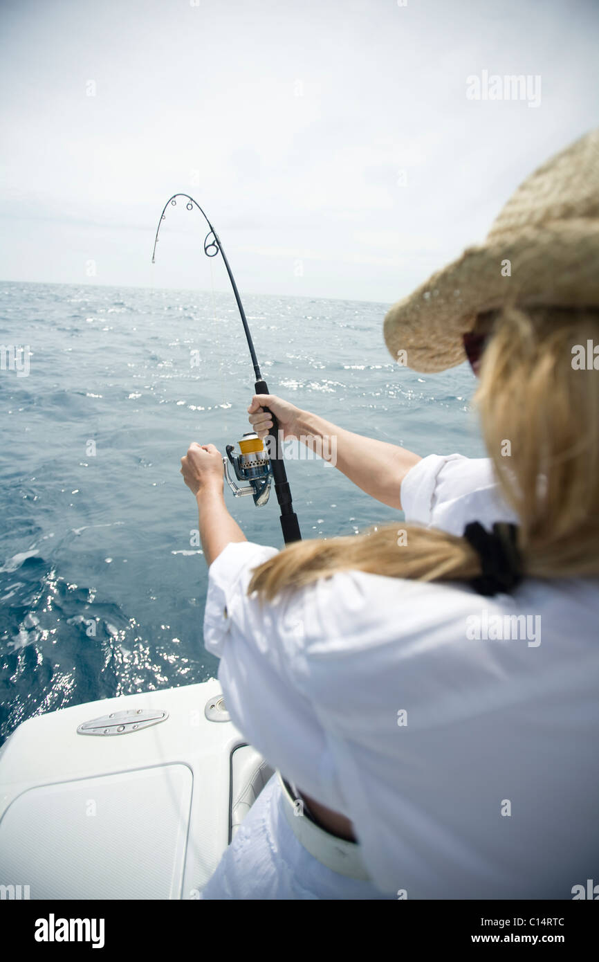 A blonde woman in a straw hat reels in a fish with blue water in the distance. Stock Photo
