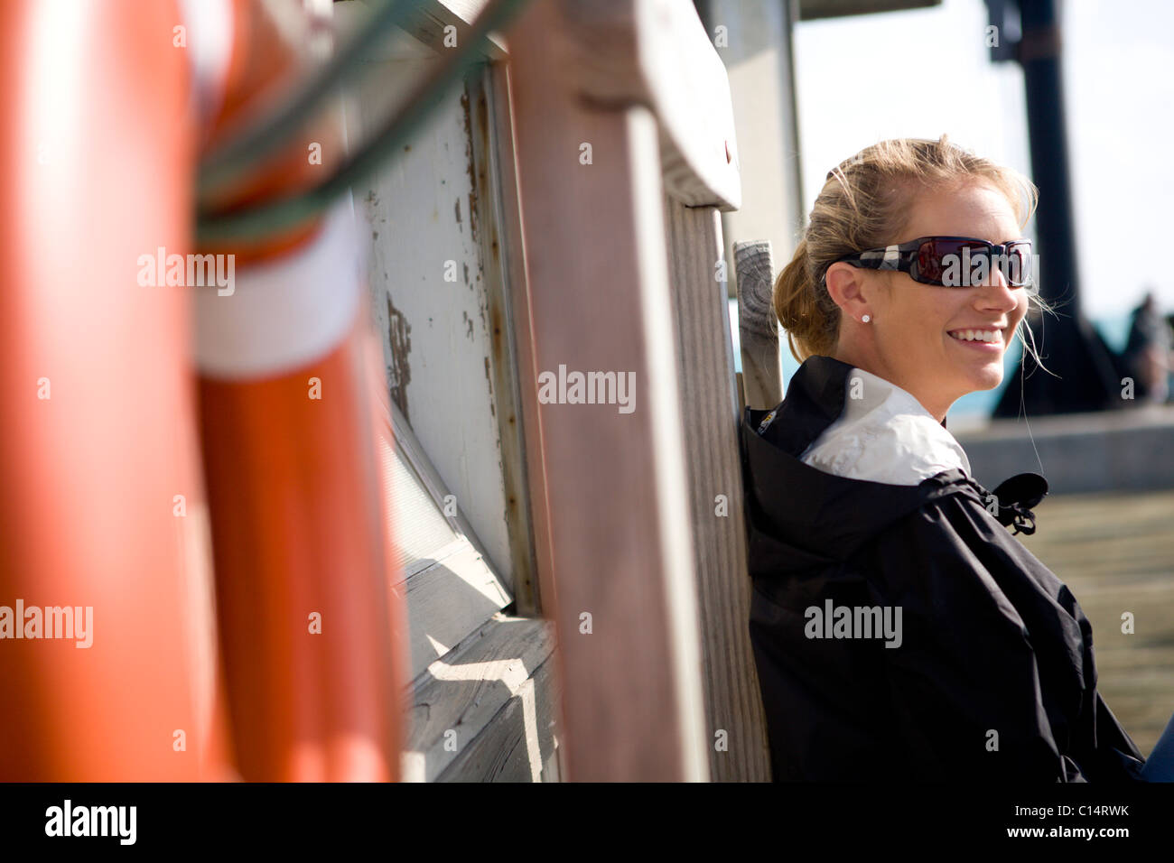 A mid adult woman sits against a dock with an orange life preserver in the foreground. Stock Photo