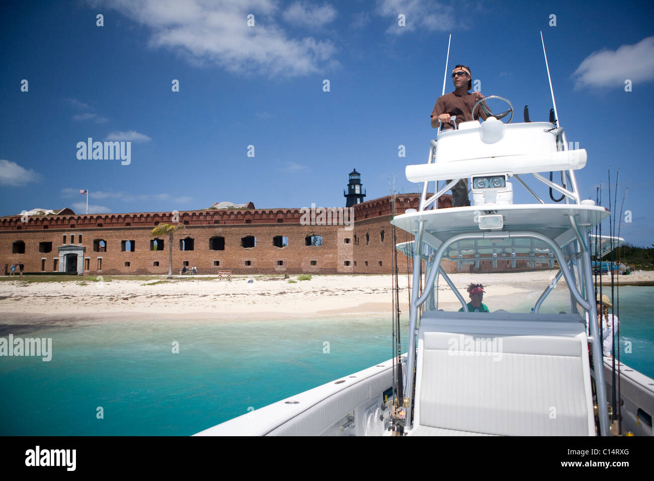 A fisherman steers his boat from the fly bridge pulling away from an old fortress with a lighthouse. Stock Photo
