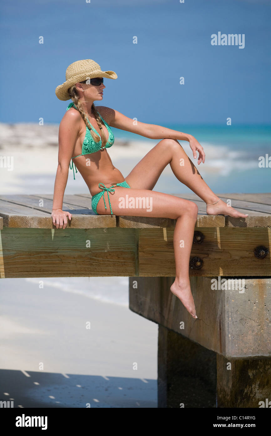 a blonde woman in pigtails is wearing a green bikini and straw hat while sitting on a beach boardwalk. Stock Photo