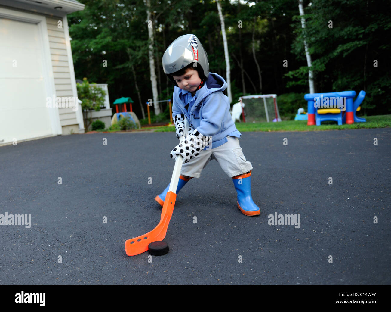 2 year old boy plays street hockey with oven mitt gloves and rubber boots Stock Photo