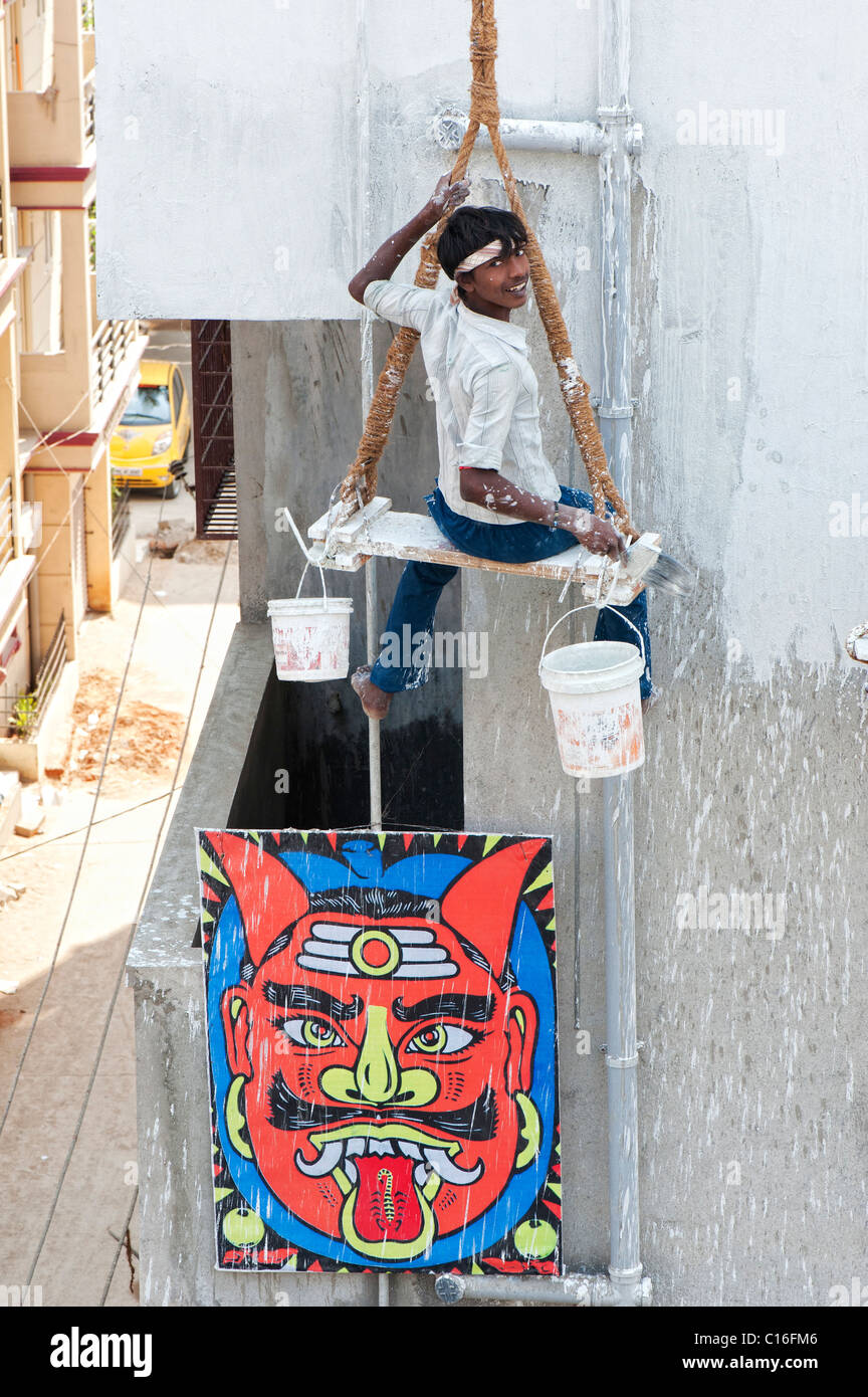 Indian painter sitting on a wooden seat hanging from ropes painting the side of an apartment complex. Puttaparthi, Andhra Pradesh, India Stock Photo