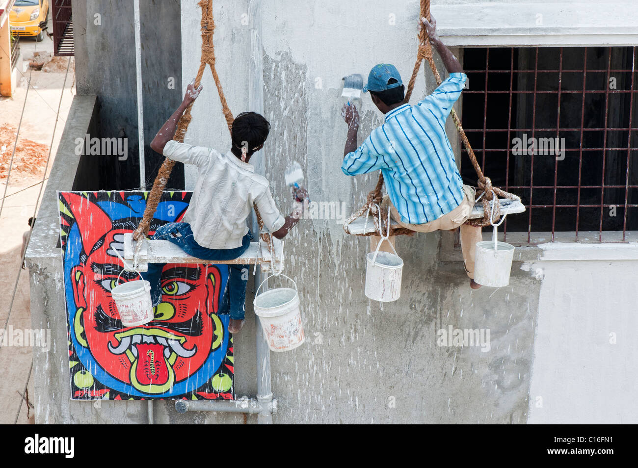Two indian painters sitting on a wooden seat hanging from ropes painting the side of an apartment complex. Puttaparthi, Andhra Pradesh, India Stock Photo