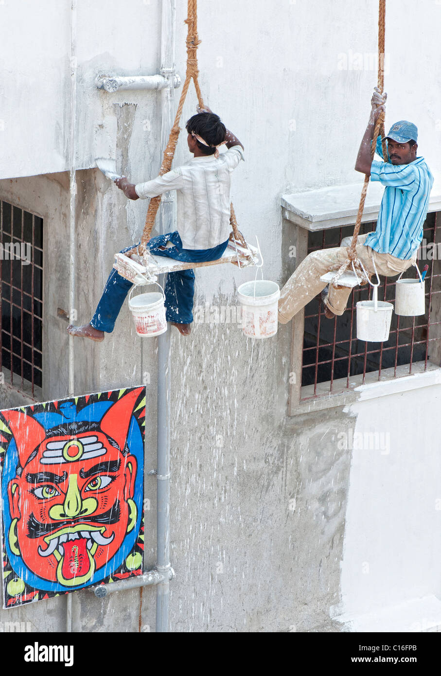 Two indian painters sitting on a wooden seat hanging from ropes painting the side of an apartment complex. Puttaparthi, Andhra Pradesh, India Stock Photo