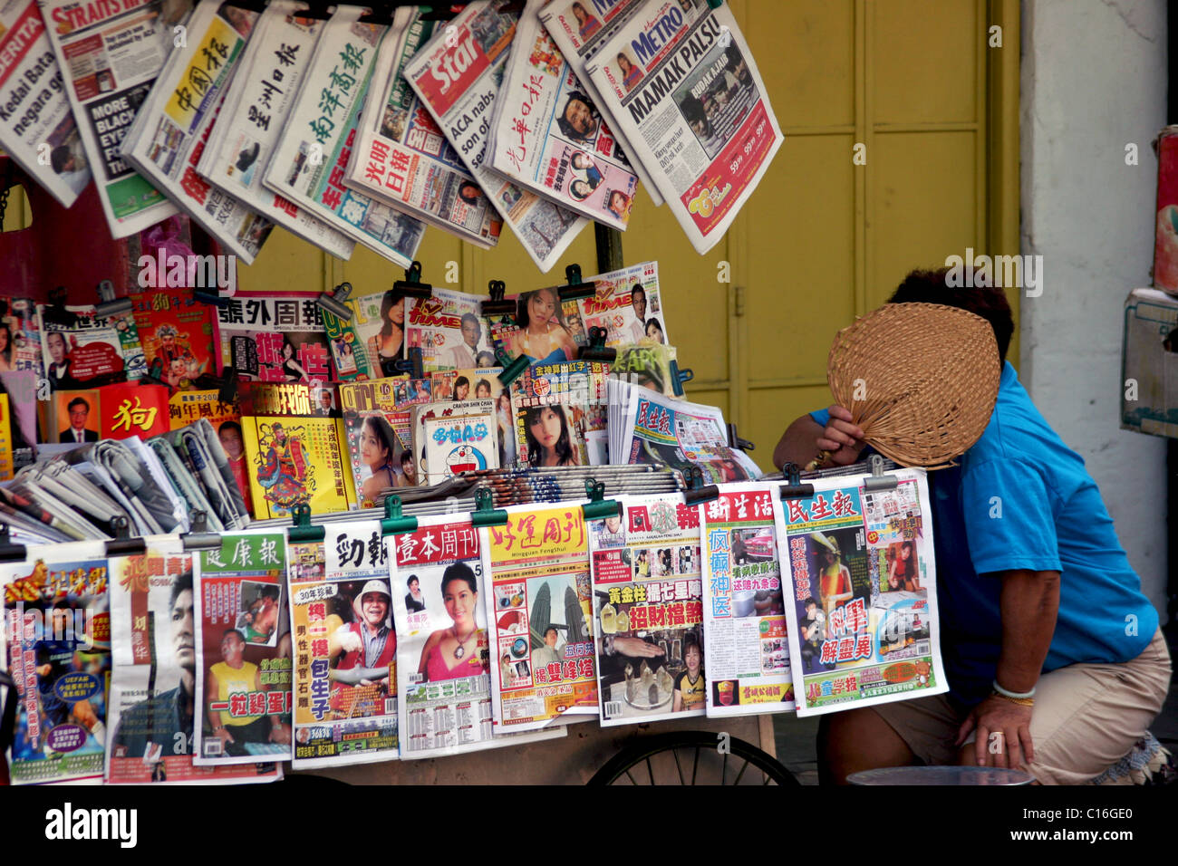 A woman is holding a fan in front of her face near magazines with pictures of faces at a news stand in Georgetown, Penang, Malay Stock Photo