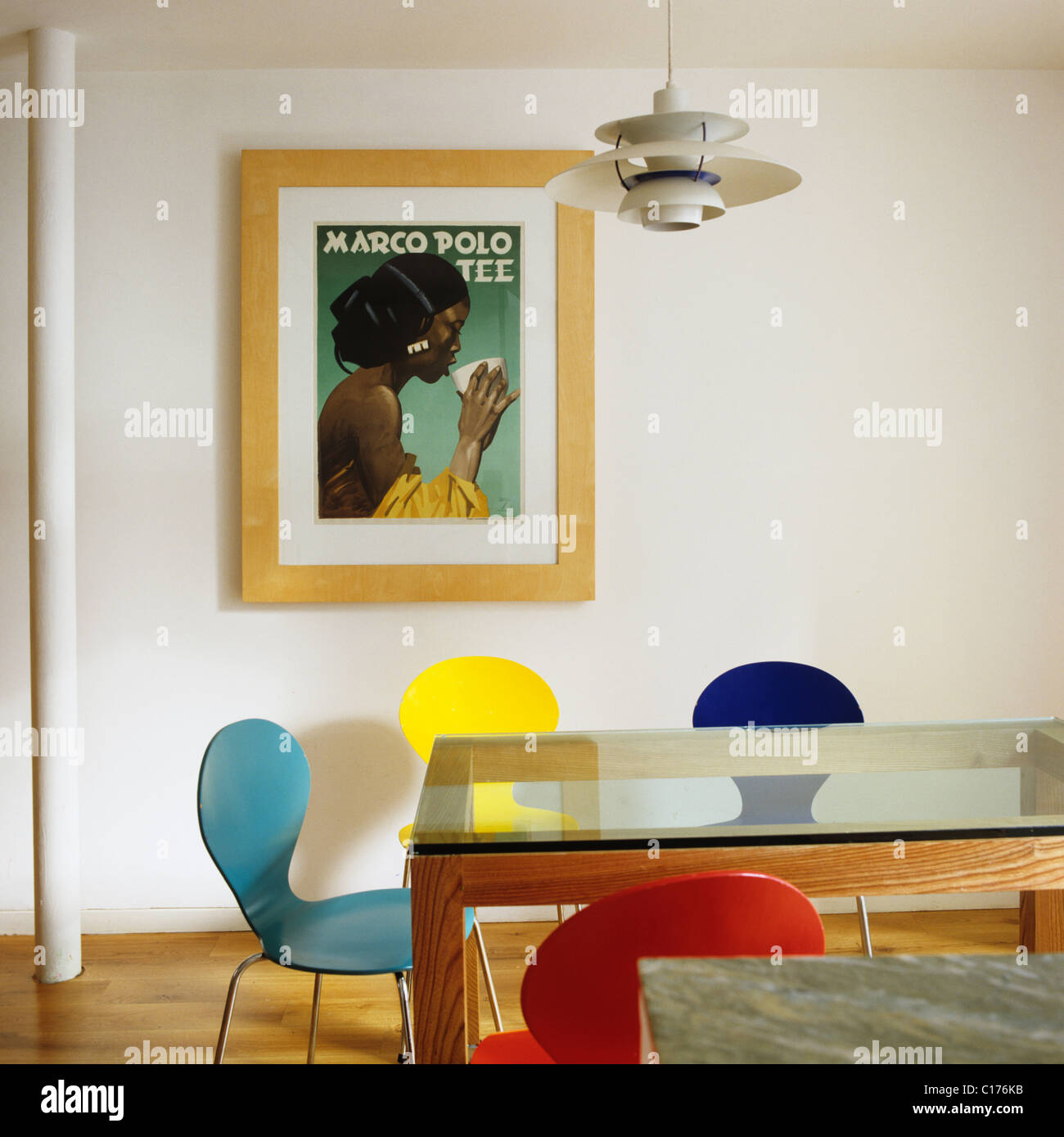 Brightly coloured butterfly chairs around a glass topped dining table in room with Ludwig Hohlwein 'Marco Polo Tee' poster Stock Photo