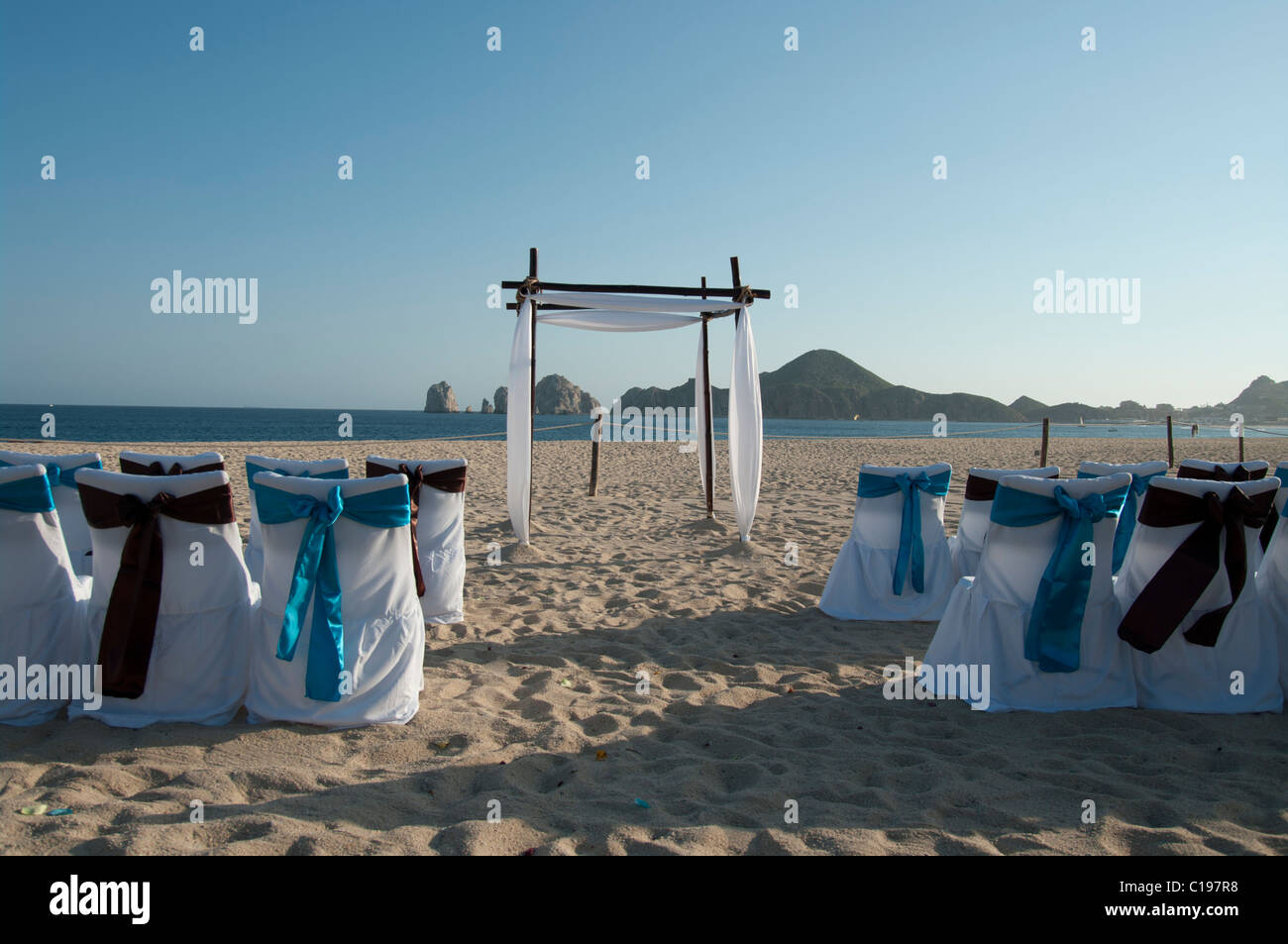 A deserted beach set up for a wedding ceremony.  The chair covers are white with blue and brown bows. An arch awaits the couple. Stock Photo