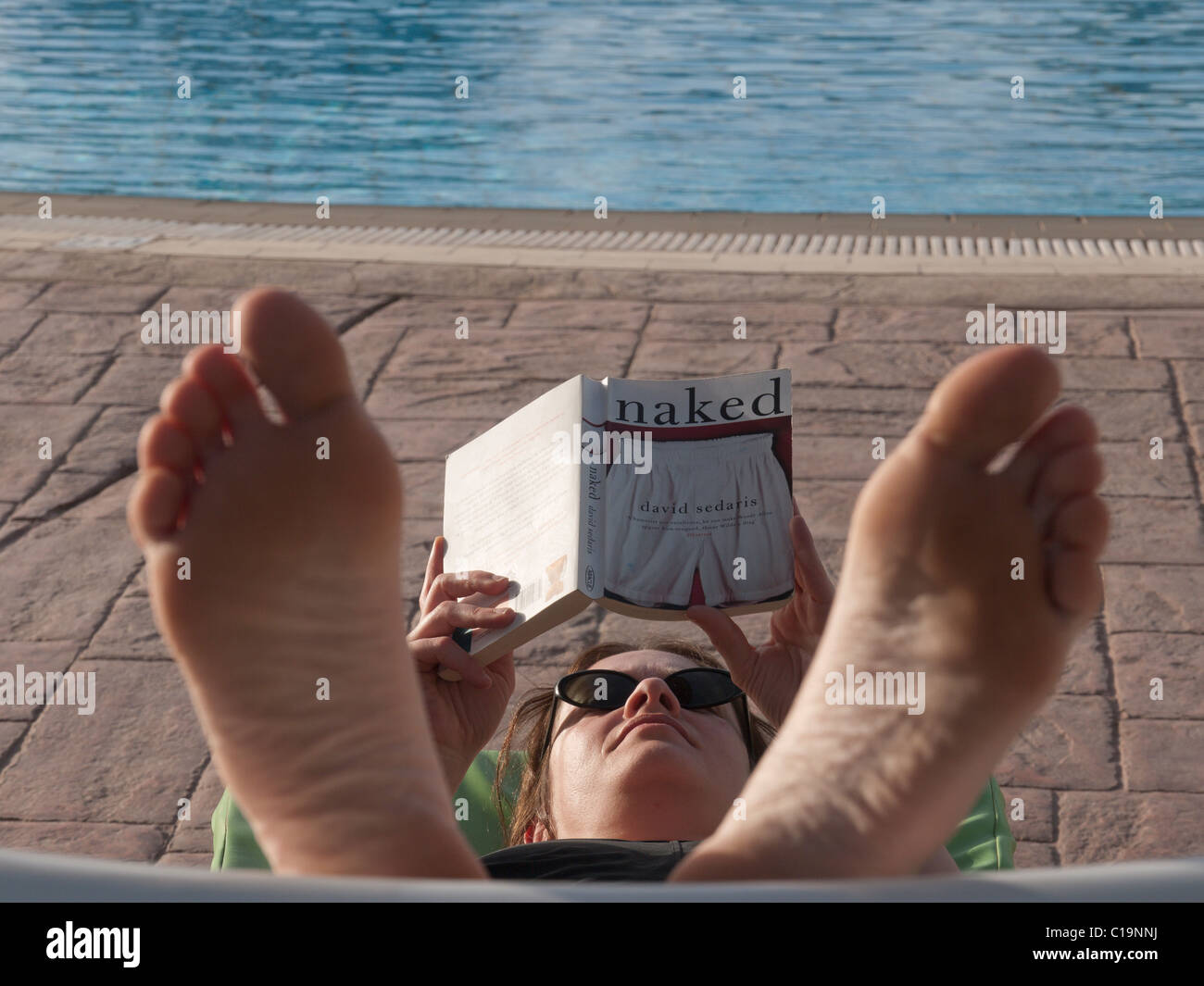a young woman with feet up relaxes by reading beside swimming pool in