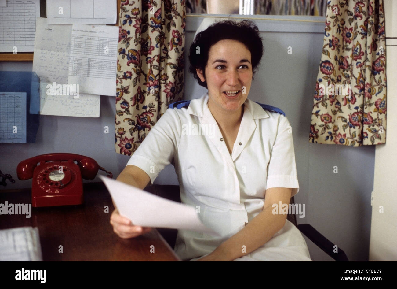 A young NHS nurse smiling in a white uniform sitting at a desk next to a red telephone holding a letter in a hospital administration office in the 1970s 1980s in Bronglais hospital Aberystwyth Wales UK   KATHY DEWITT Stock Photo