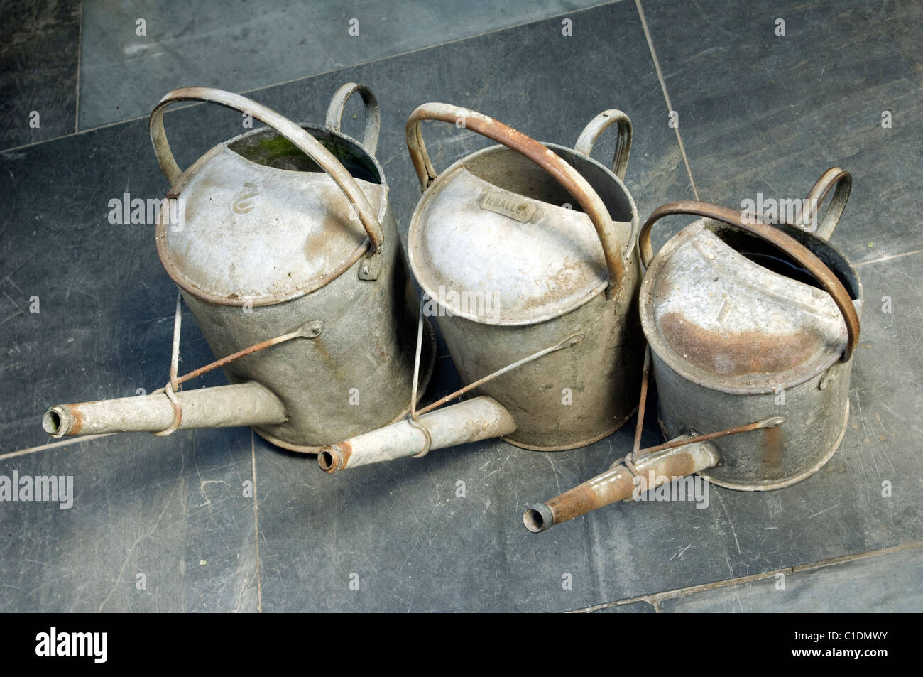 Three galvanised gardeners Watering Cans, a One Gallon, One and a Half Gallon and a Two Gallon on a slate floor. Stock Photo