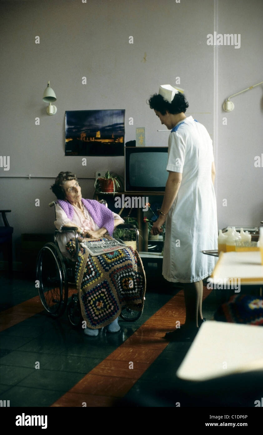 An elderly woman patient with a crocheted shawl sitting in a wheelchair talking to a nurse in an NHS dayroom at the Bronglais hospital in 1980s 1980 Wales UK  KATHY DEWITT Stock Photo