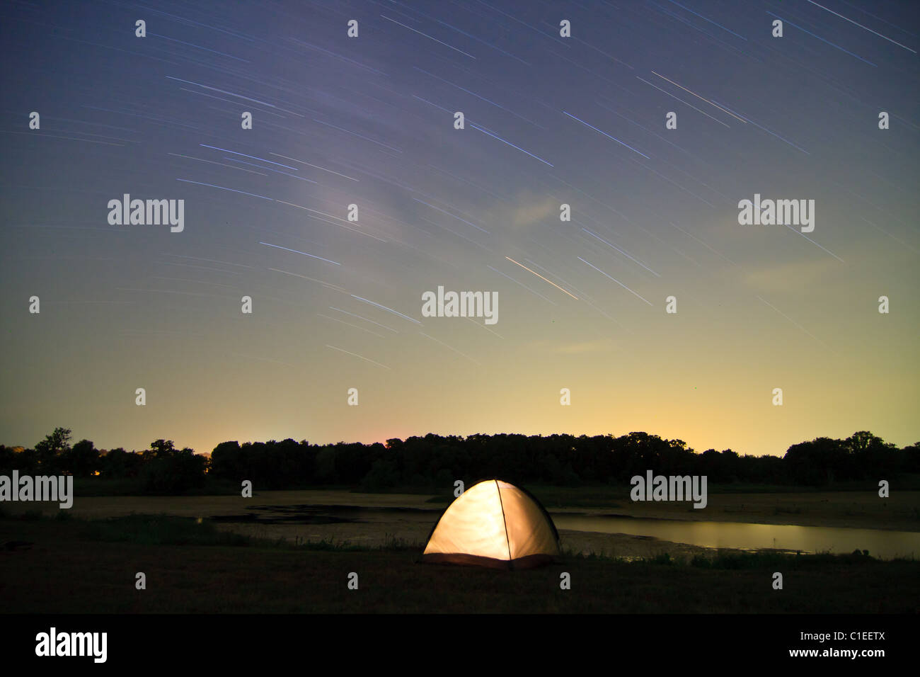 Lone illuminated tent camped at night in front of a lake with star trails overhead at Hubbard City Lakes in Hubbard, Texas, USA. Stock Photo