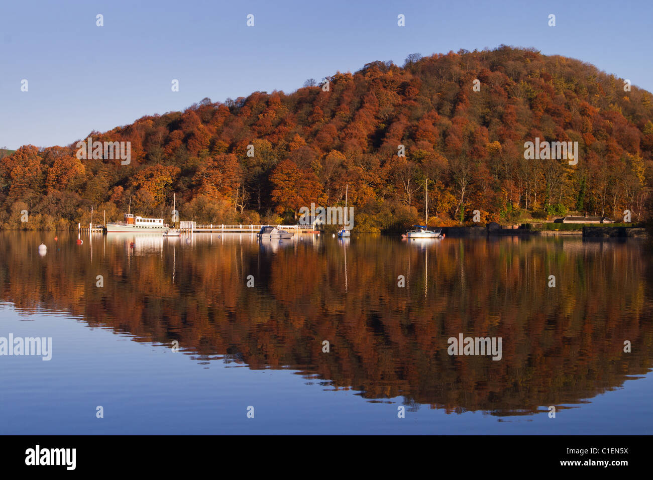 Boats, pier and autumn trees reflected in lake water. Ullswater, Lake District, England. Stock Photo