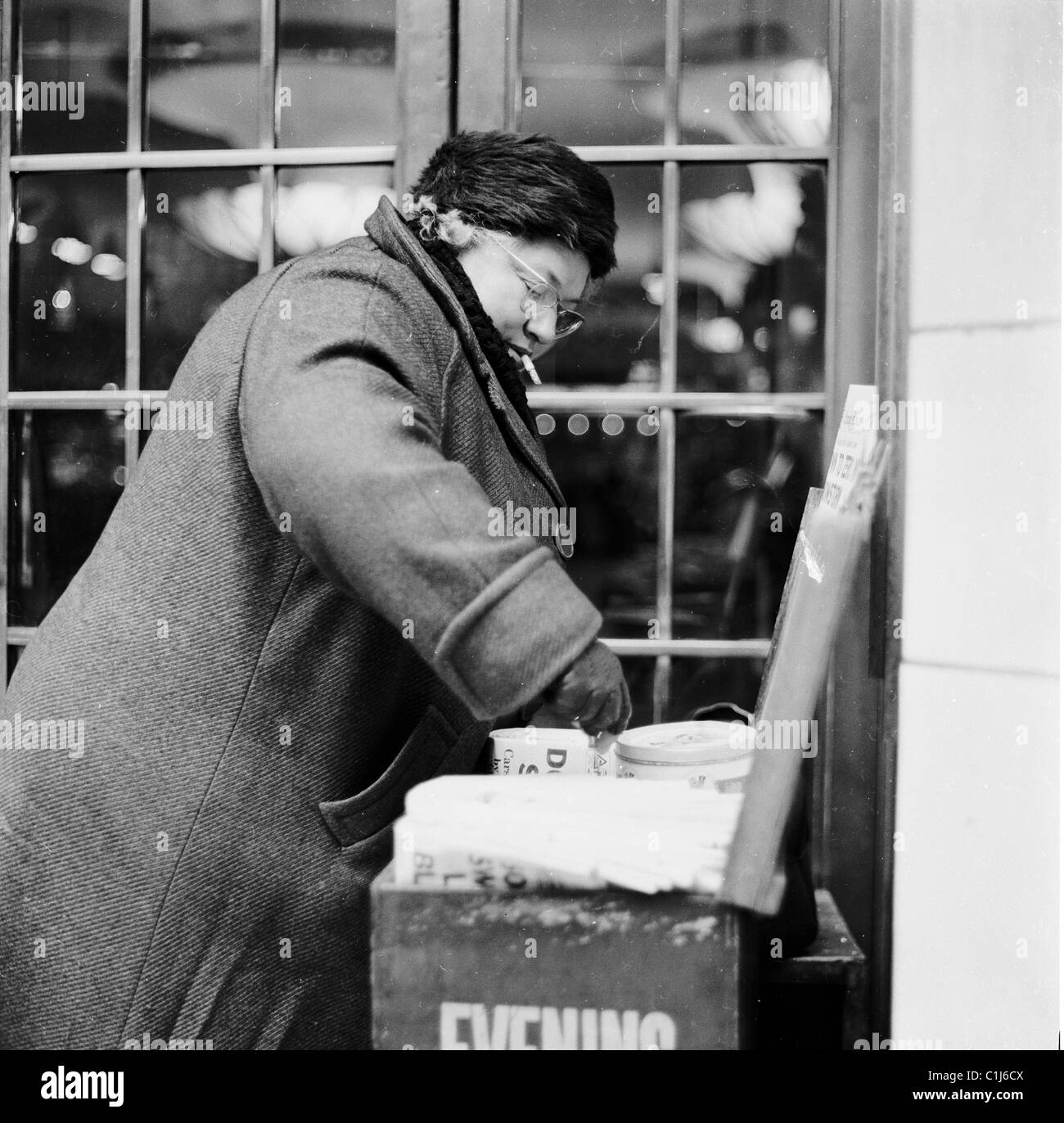 1950s, winter time and a female evening newspaper seller wearing a thick coat, hat, gloves and smoking, sorts out her change, London, England, UK. Stock Photo