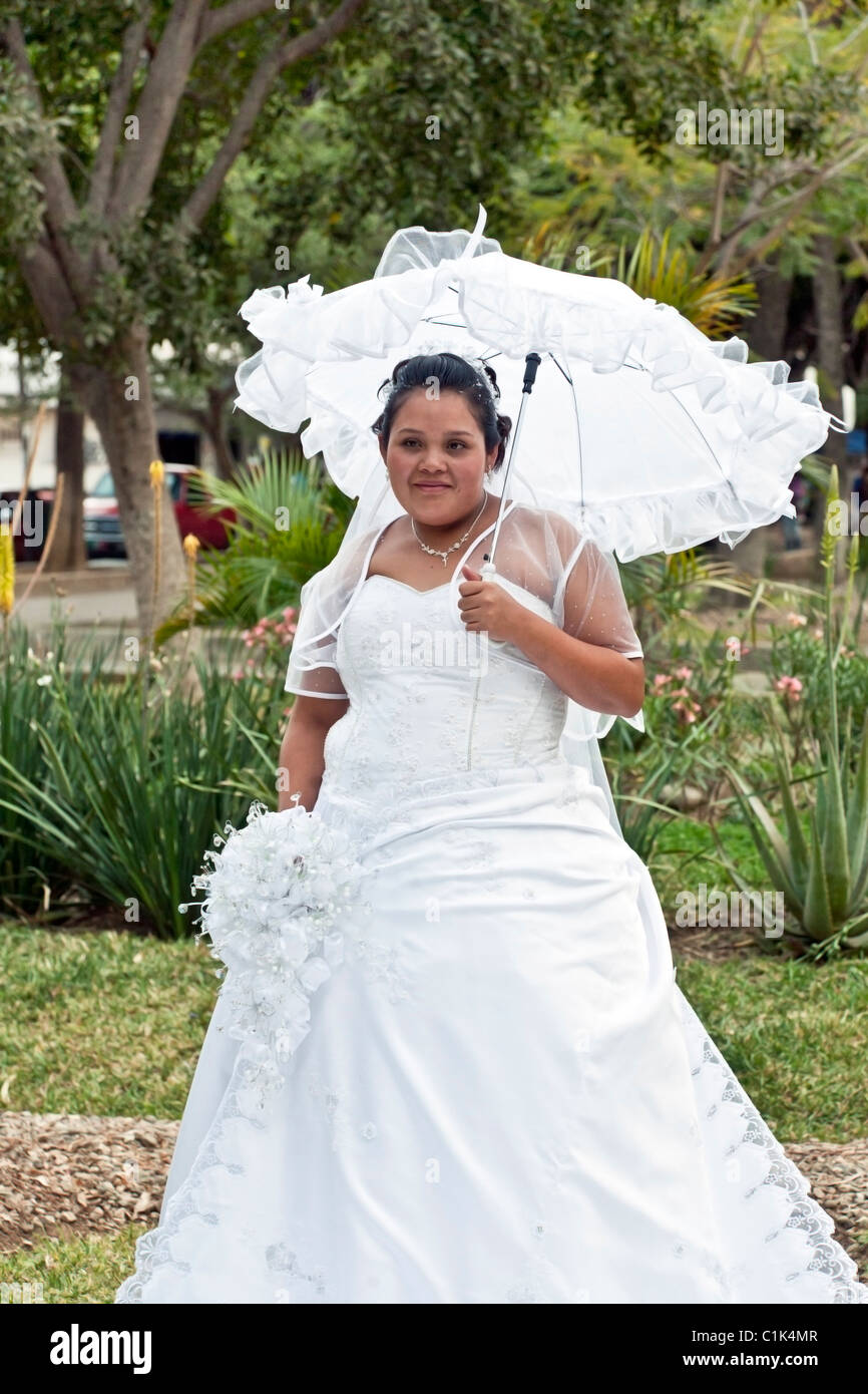 plump smiling bride poses in her wedding dress under shade of frilly white parasol in Benito Juarez Park garden Oaxaca Mexico Stock Photo