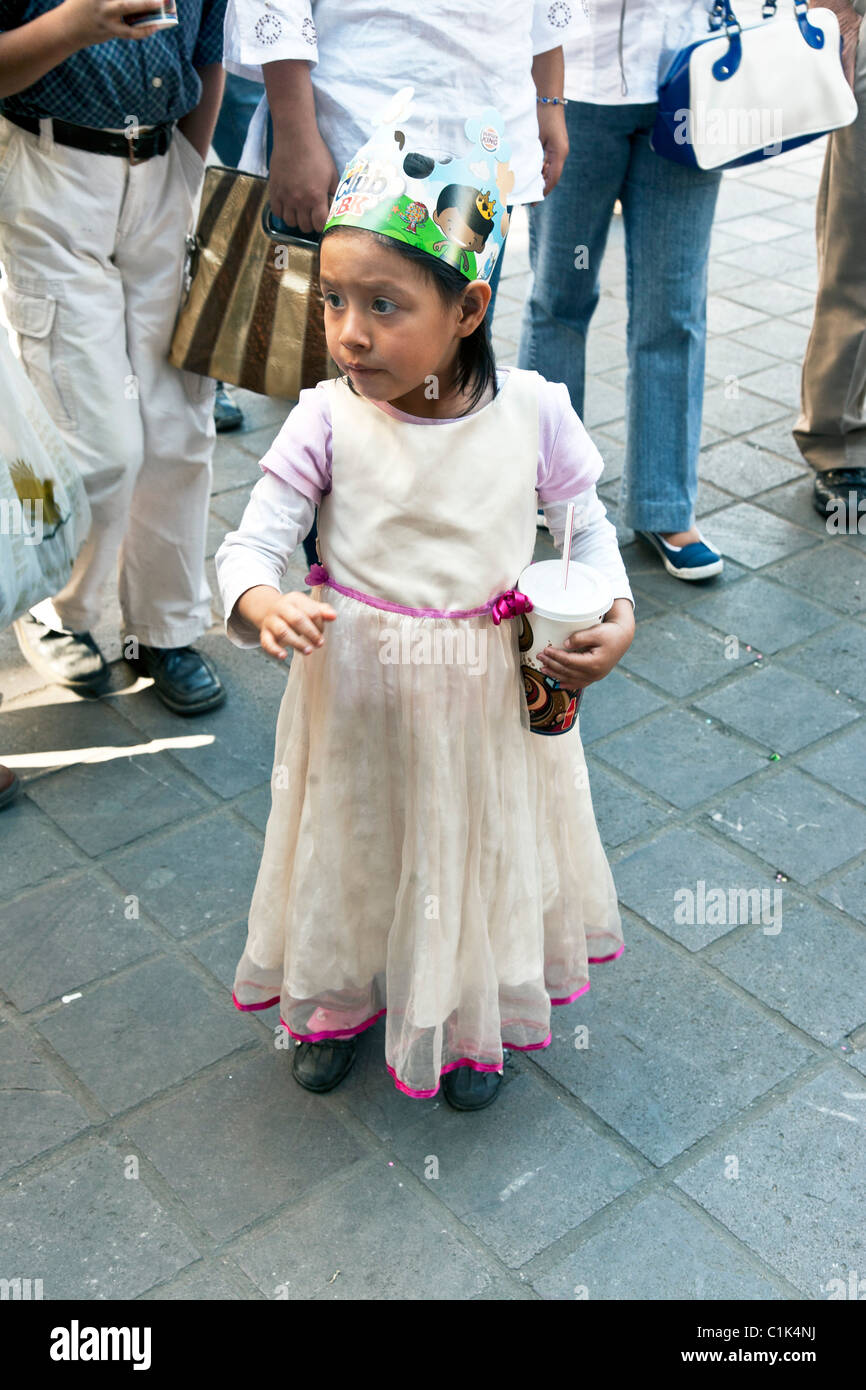 solemn little Mexican girl wearing chiffon skirt & Burger King cardboard crown stands in Oaxaca Zocalo Stock Photo