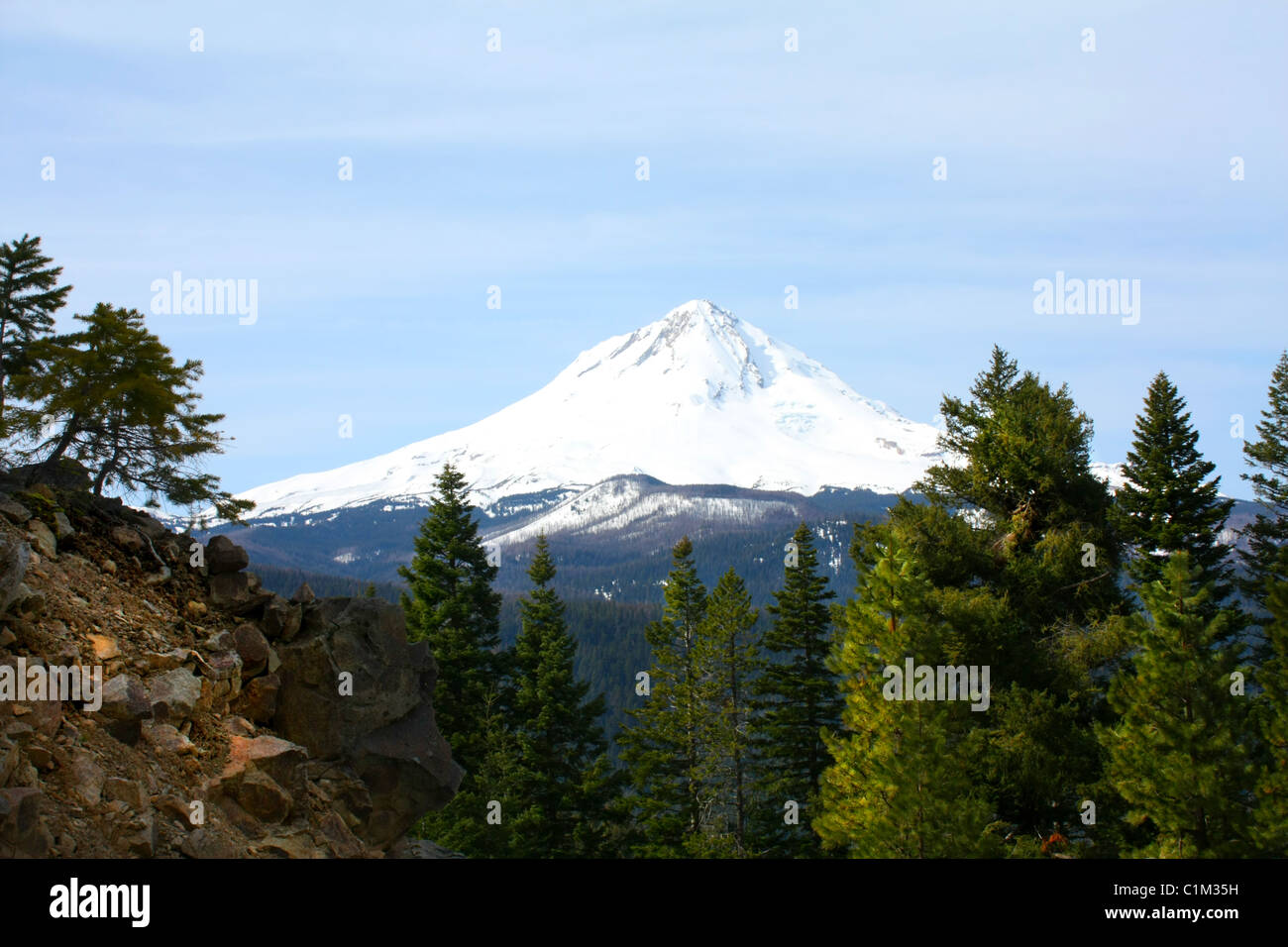 40,257.07614 A distant snow-covered mountain - Mt Hood (11,240 ft tall) - peaks over a tree covered rocky ridge top, against a soft blue sky. Stock Photo