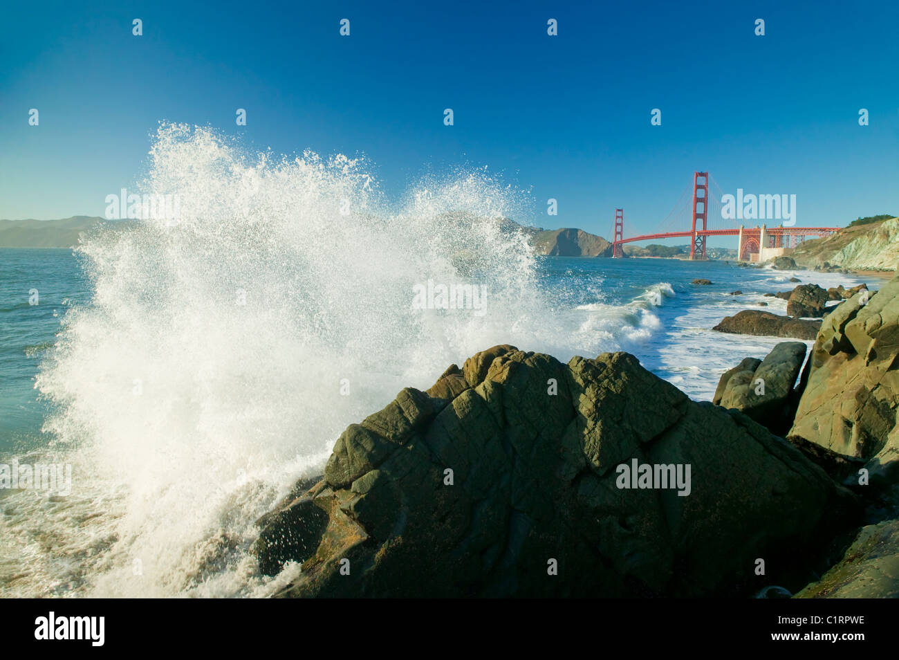 A large ocean wave crashing into rock at Baker Beach, Golden Gate Bridge in background Stock Photo