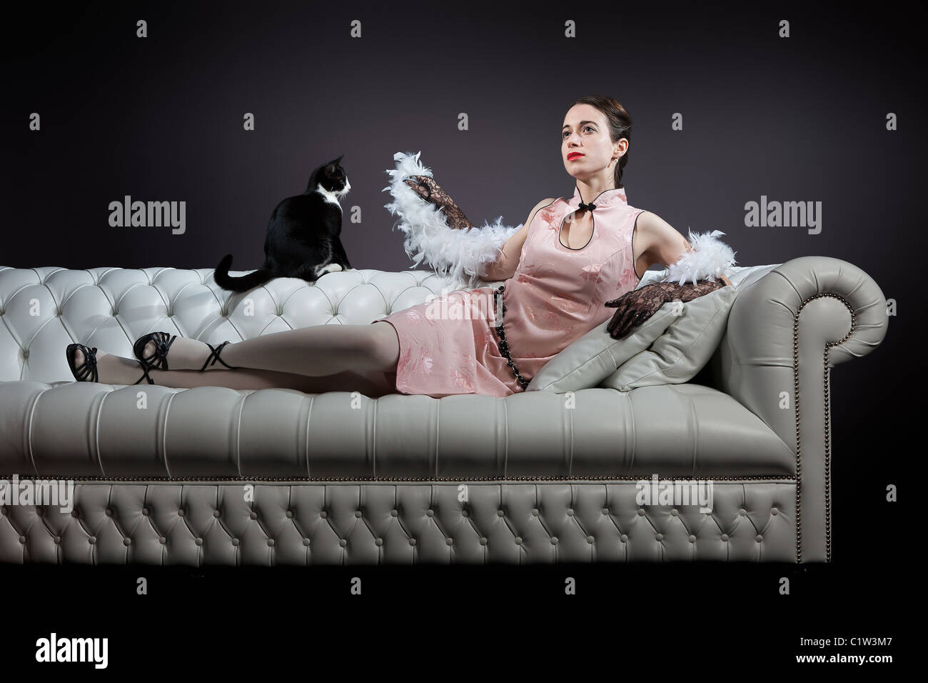 An elegant vintage lady is lying on a sofa, with a kitten looking at her feather boa. Stock Photo