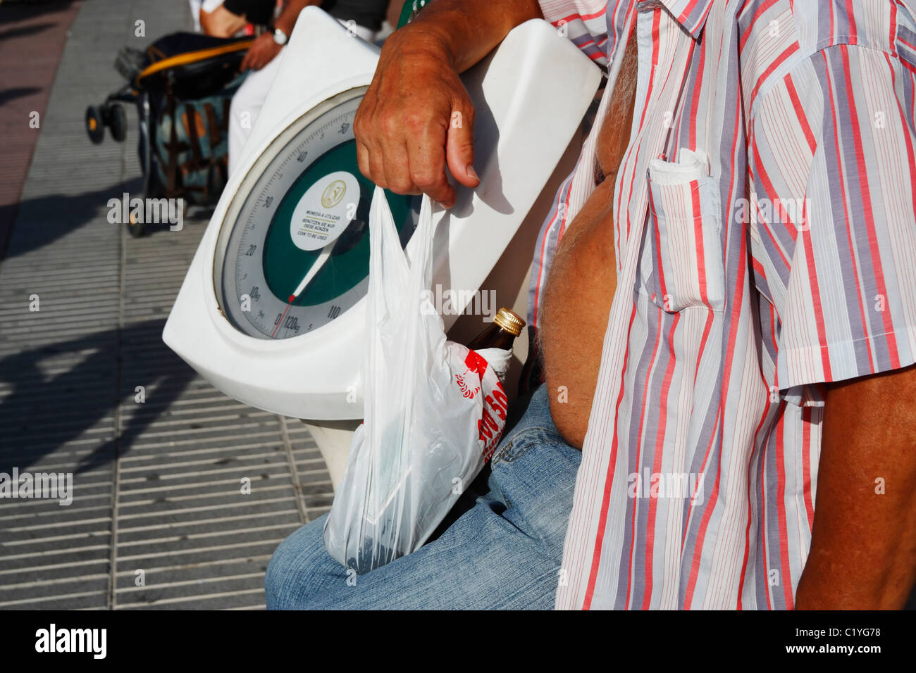 Man with bottle of beer in plastic bag sitting on wall next to coin operated weigh machine in Spain Stock Photo