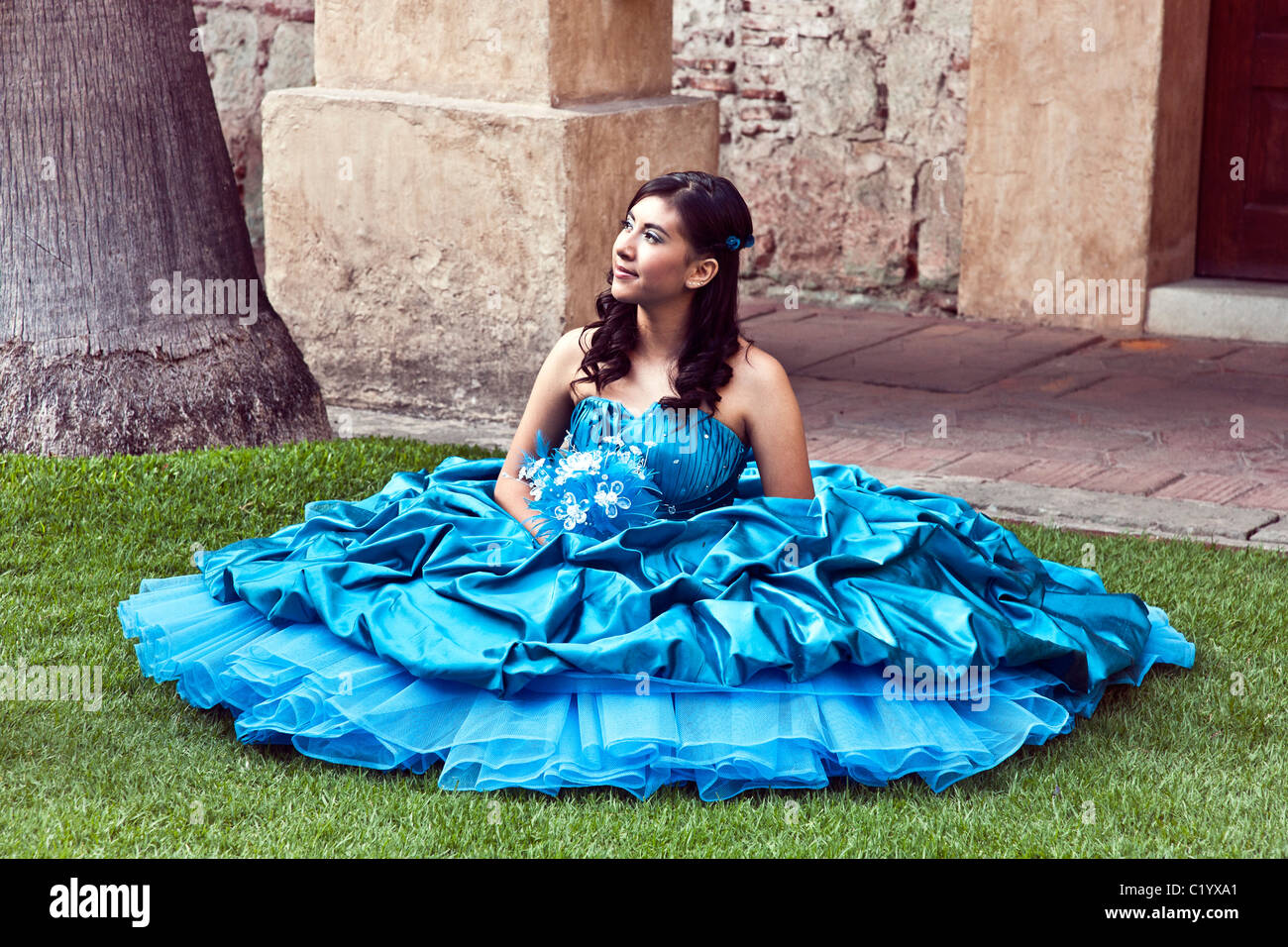 pretty teen Mexican girl in brilliant turquoise strapless gown poses for her quinceanera portrait seated on grass Oaxaca Mexico Stock Photo