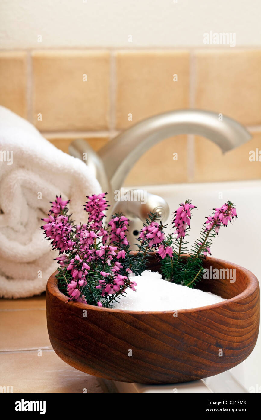 Pretty heather flowers in a bowl of Epsom salts on a bath tub edge ready to bathe. Epsom salts with purple heather flowers in a wood bowl near a tub Stock Photo