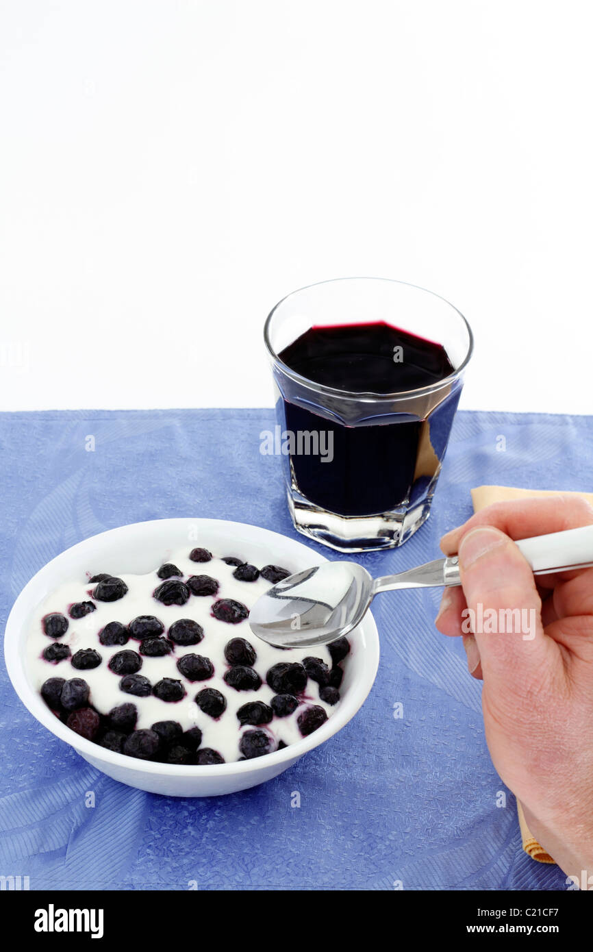 Hand with spoon about to eat a bowl of thawing frozen blueberries with vanilla yogurt, glass of organic blueberry juice. Blueberries, yogurt breakfast Stock Photo