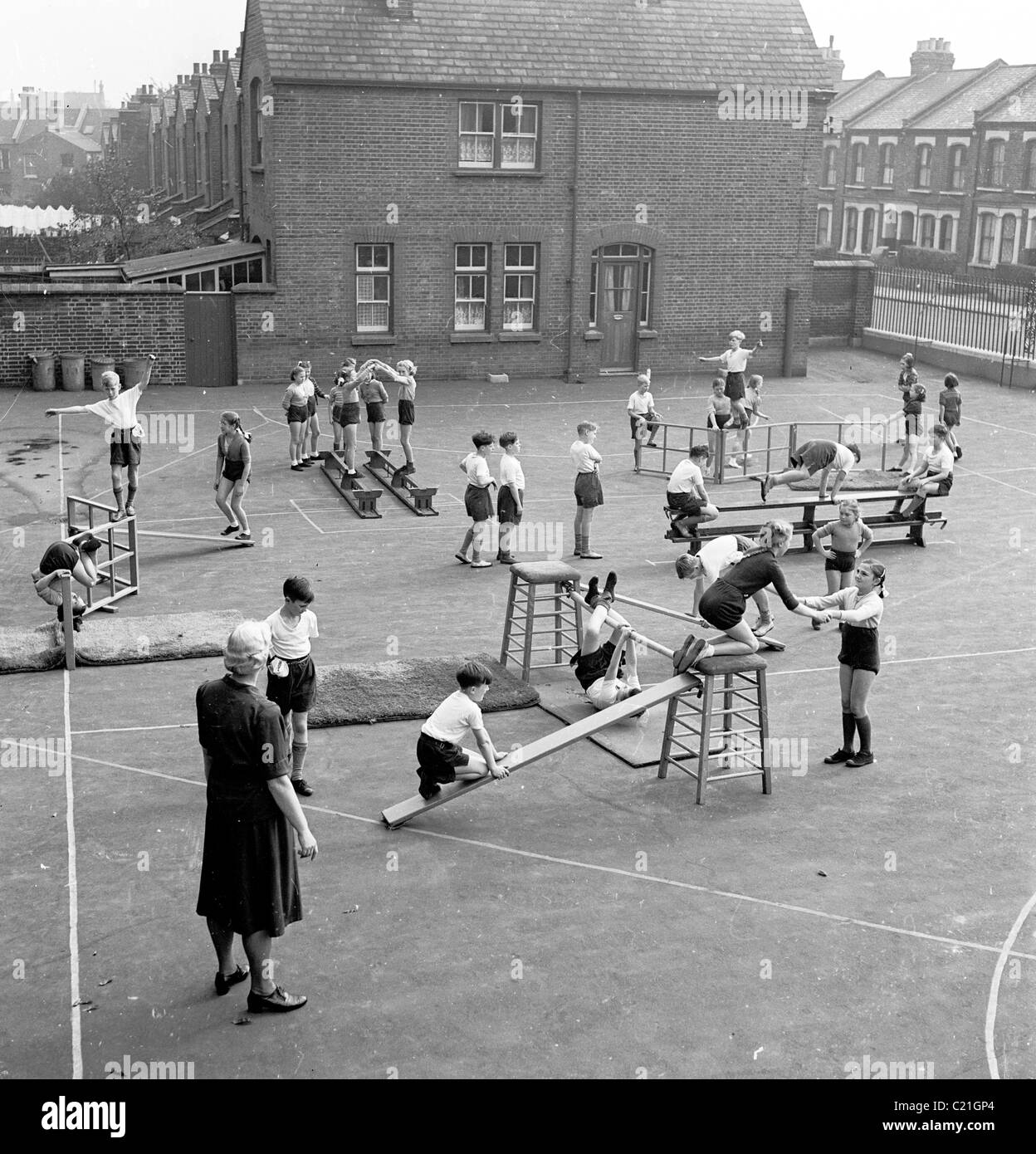 1950s, a female teacher supervises a young children doing a PE (physical education) class outside in an inner-city primary school playground, England. Stock Photo