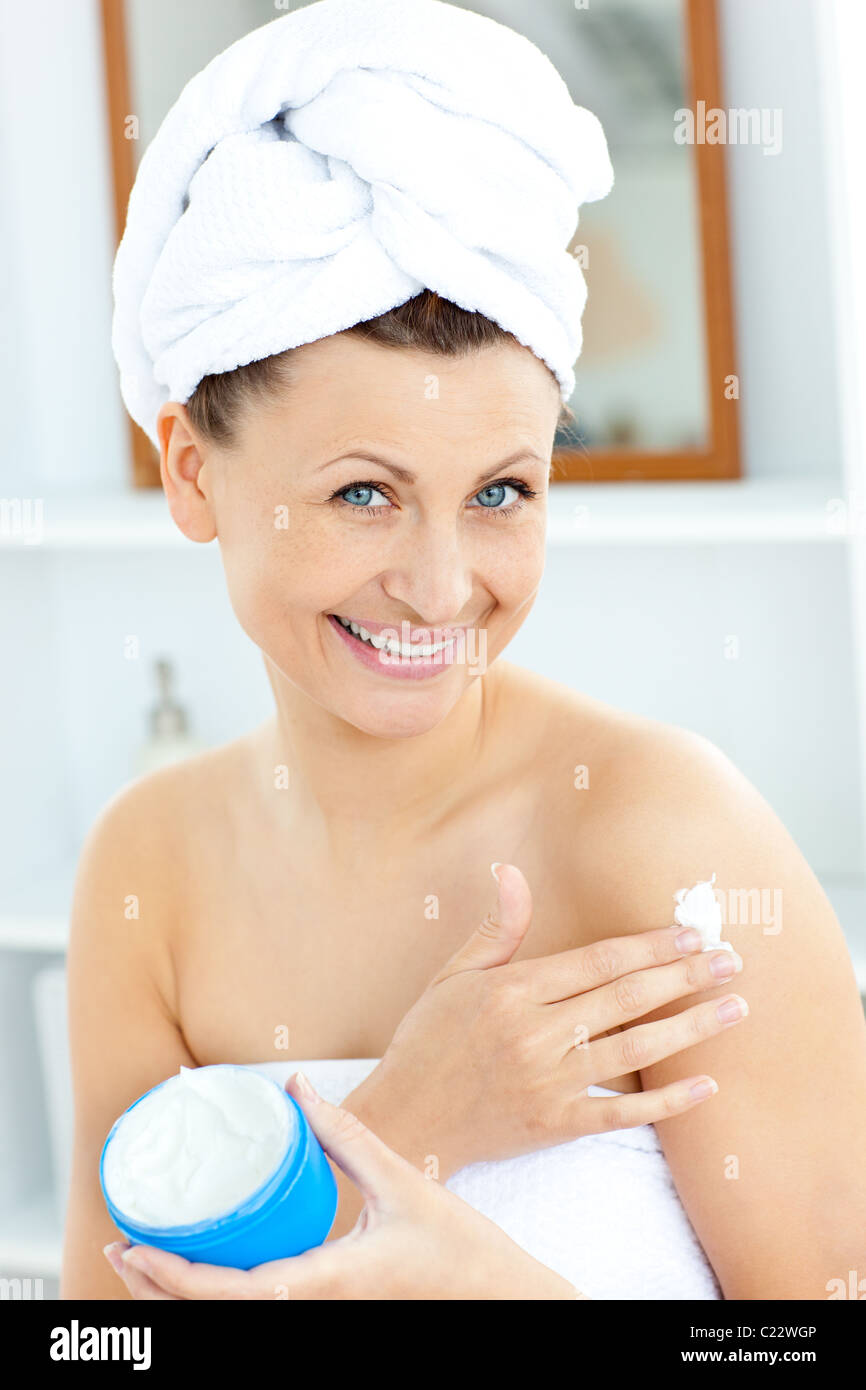 Charming young woman with a towel putting cream on her face in the