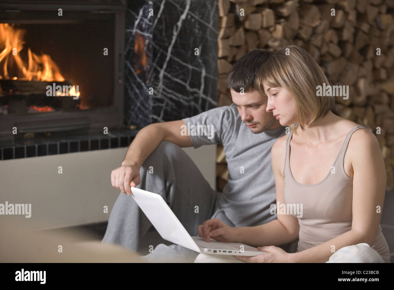 Young couple sit with laptop beside open fire Stock Photo