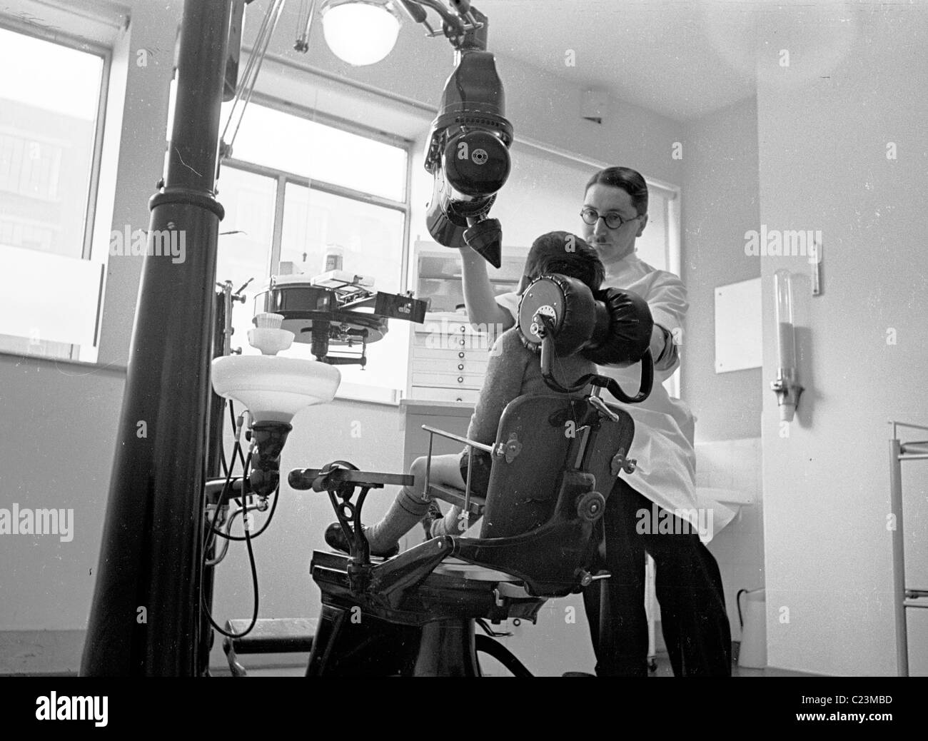 England, 1950s. A child is seen sitting in a dentists chair, surrounded by large machines, with dentist. Stock Photo