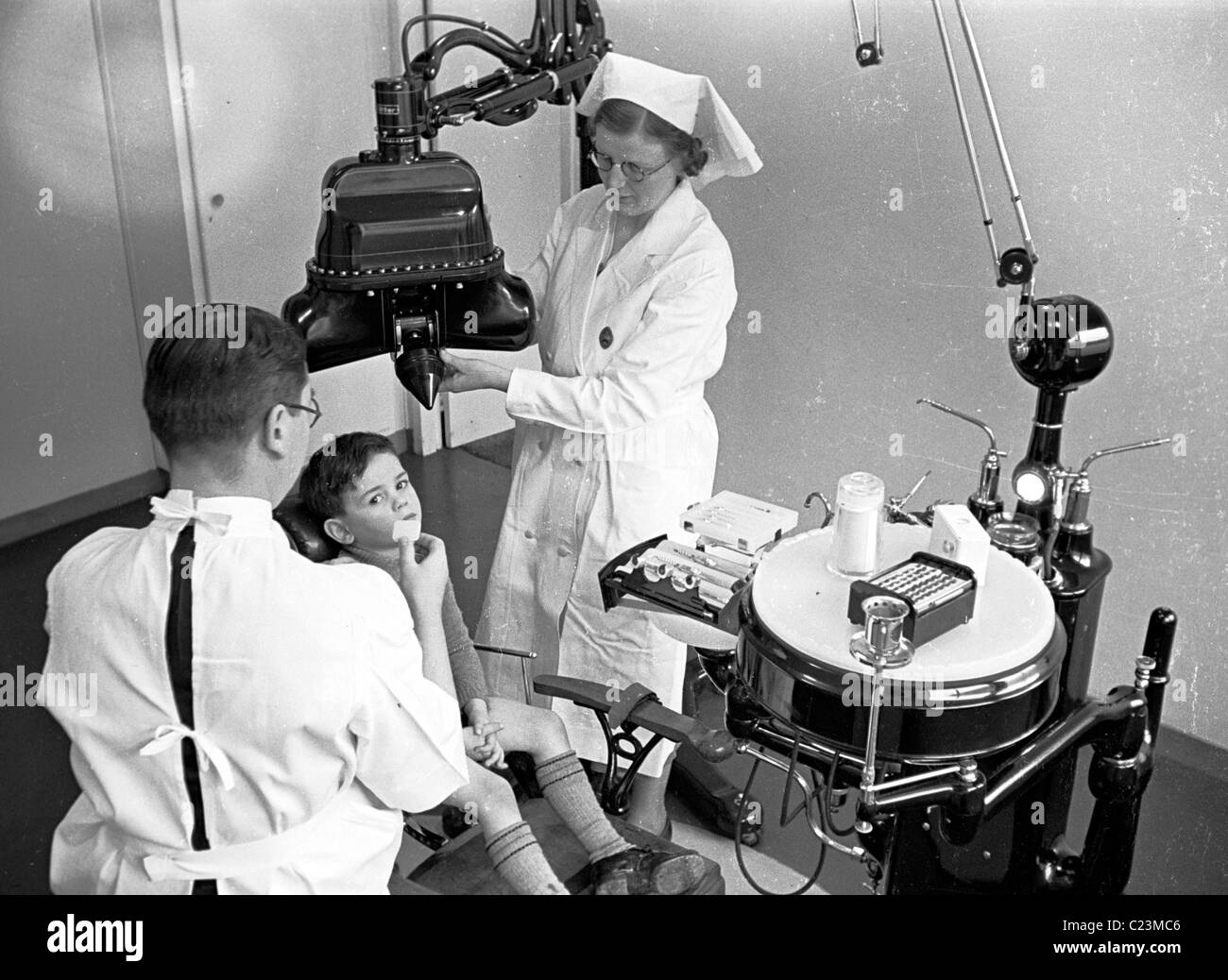 Historica 1950s. Anxious young boy sitting in a dentist's chair beside a male dentist, with a nurse using dental equipment, possibly an x-ray machine. Stock Photo