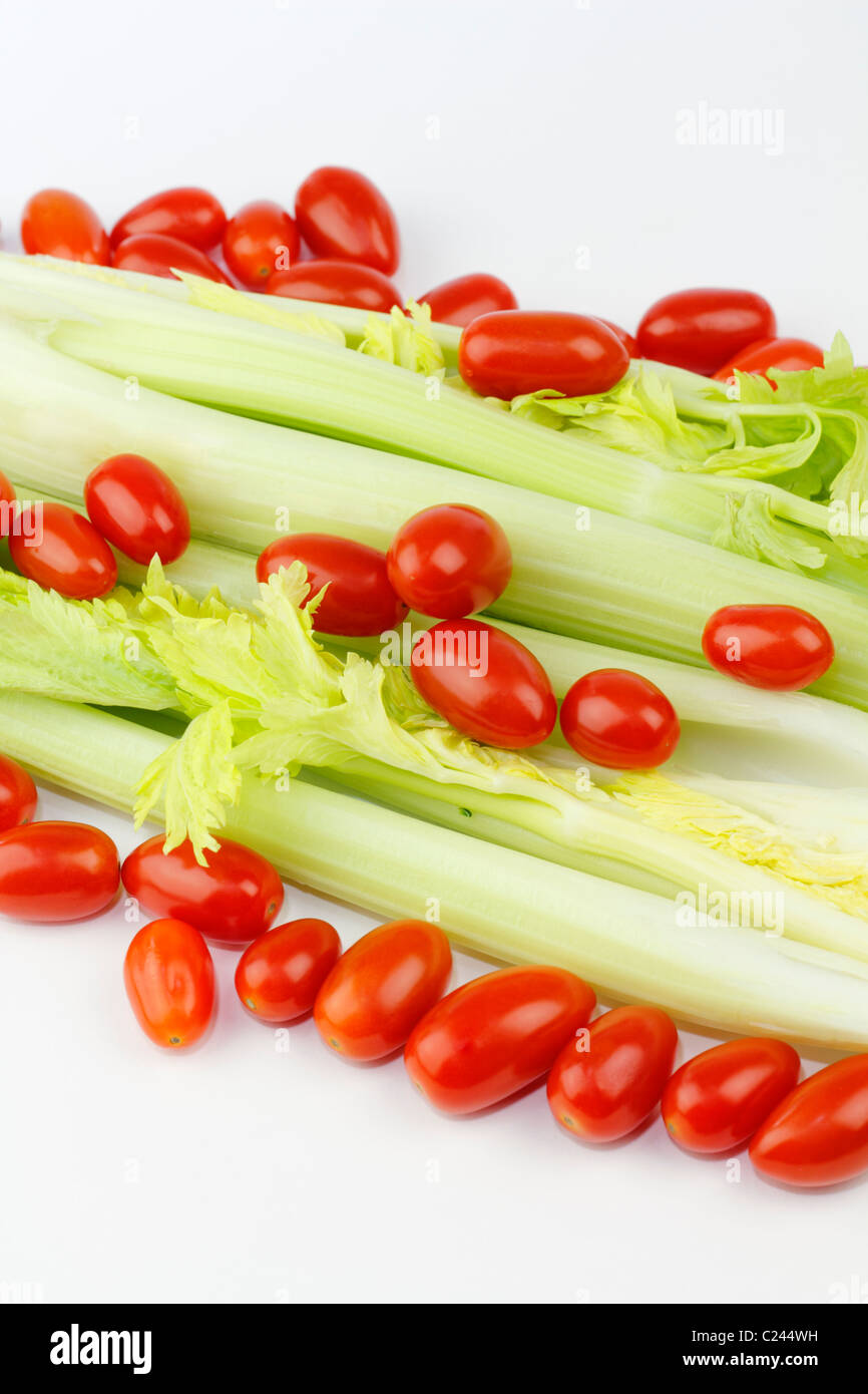 Lots of small red tomatoes with organic celery heart stalks underneath. Fresh long green vegetable and small oval red fruit mix on white at an angle. Stock Photo