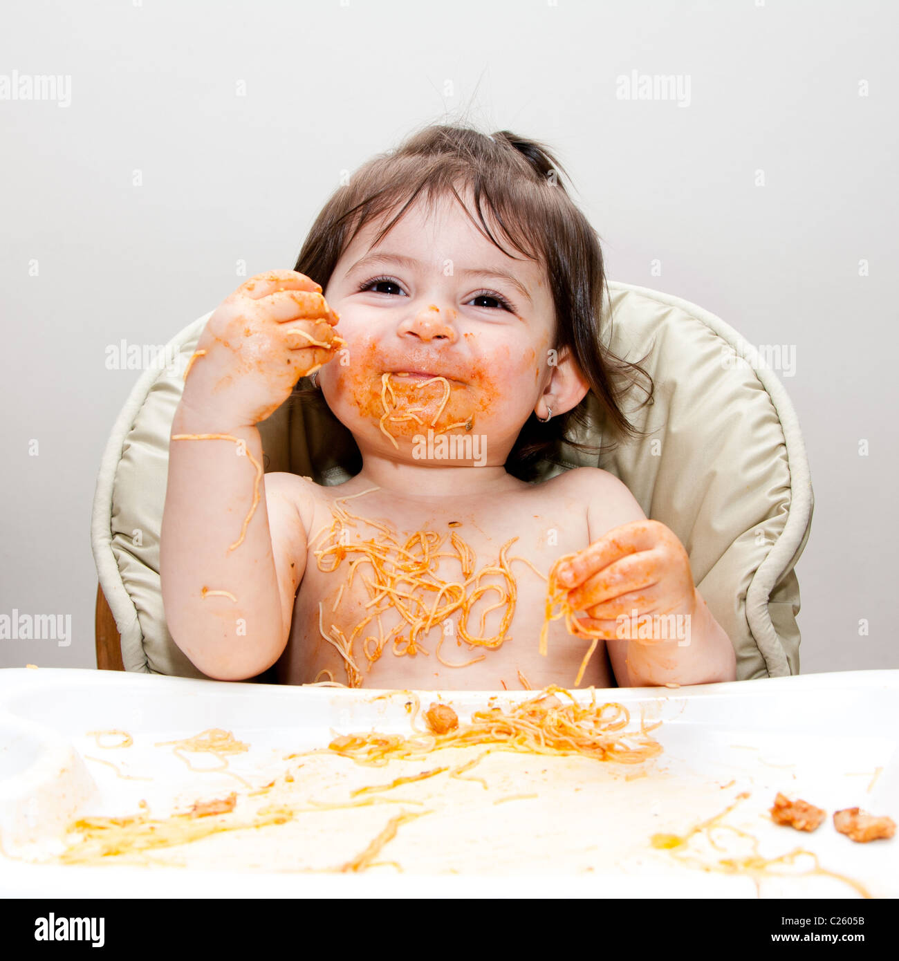 Happy smiling baby having fun eating messy covered in Spaghetti Angel Hair Pasta red marinara tomato sauce. Stock Photo