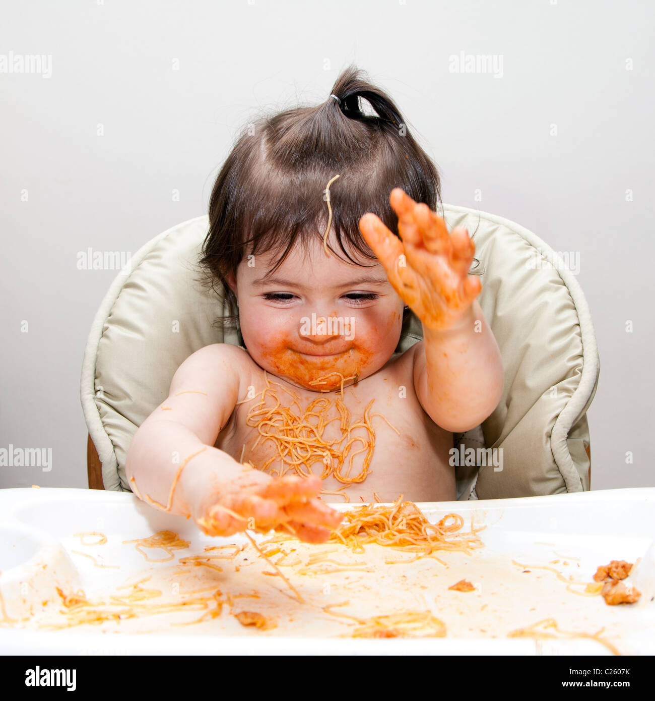 Happy baby having fun eating messy slapping hands covered in Spaghetti Angel Hair Pasta red marinara tomato sauce. Stock Photo