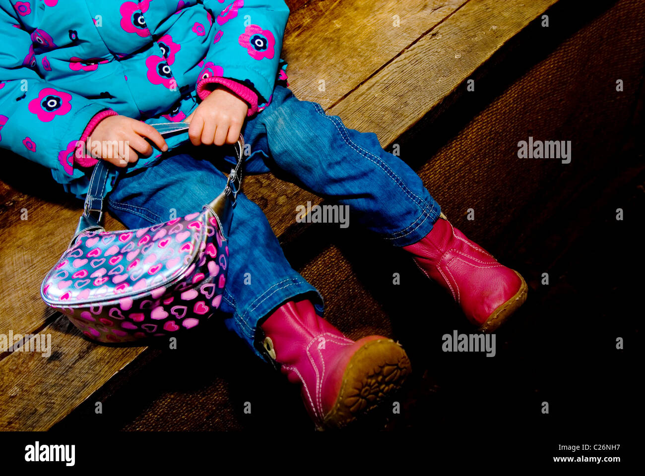Child sitting on a wooden platform with handbag wearing a blue coat with pink flowers and jeans. Stock Photo