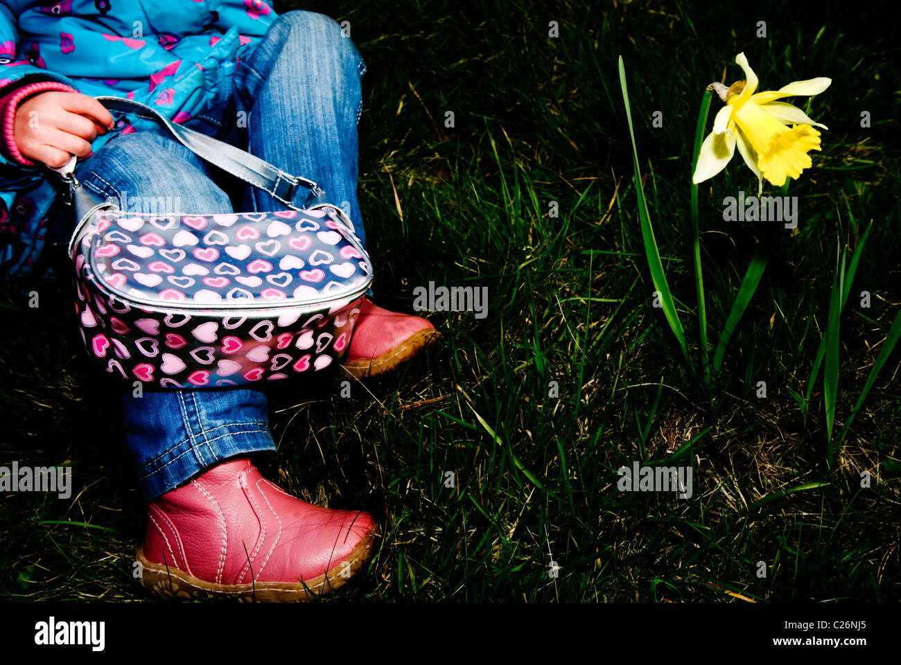 Girl sat on the grass holding a handbag looking at a daffodil Stock Photo