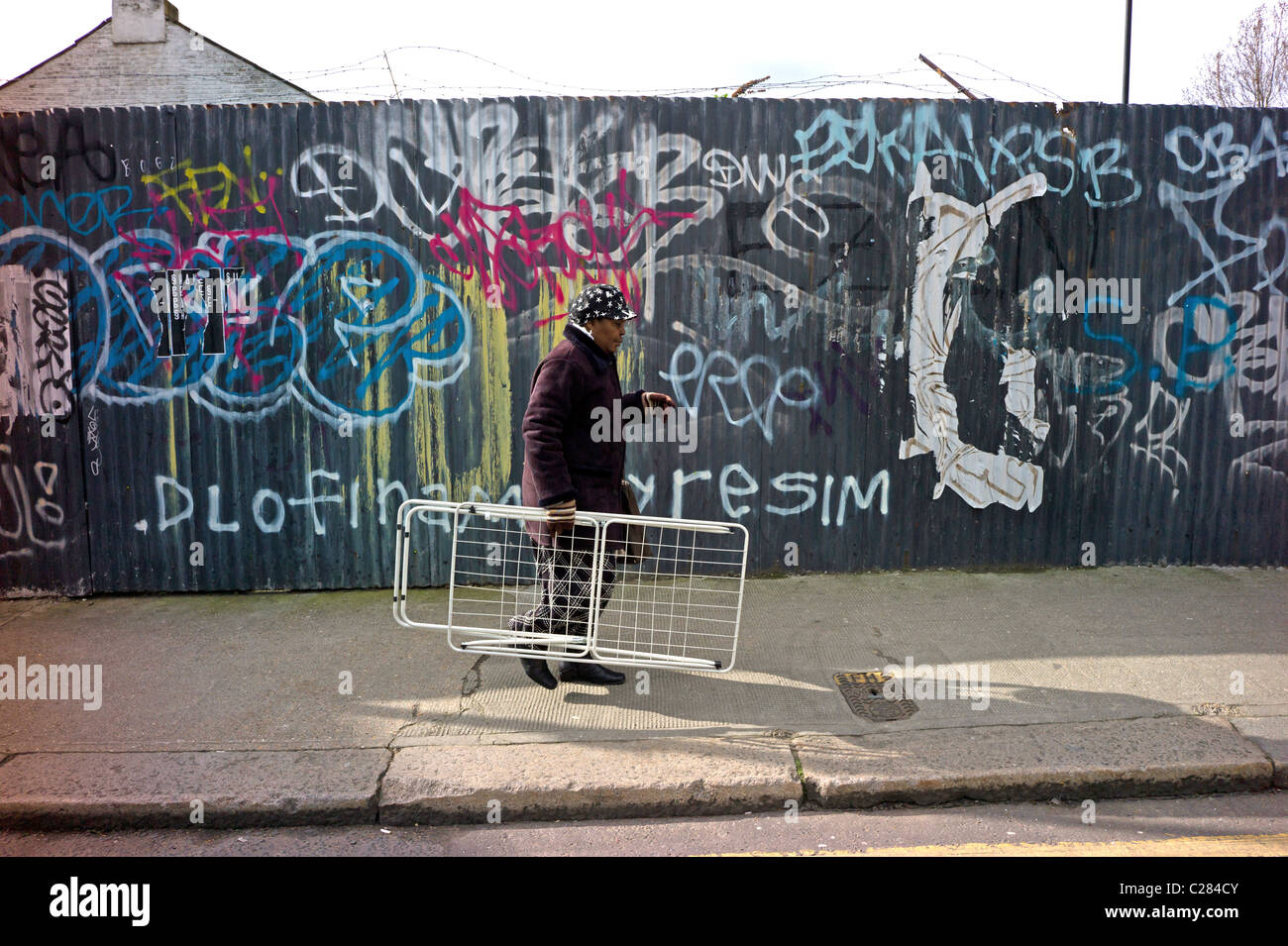 An elderly black Afro Caribbean woman carrying a clothes drying rack walking past a Graffiti covered hoarding Brick Lane London Stock Photo