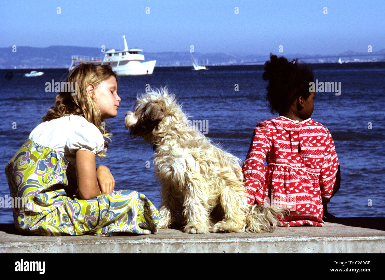 Two little girls dressed in retro sixties clothes sit on San Francisco Bay pier  playing with shaggy dog Stock Photo