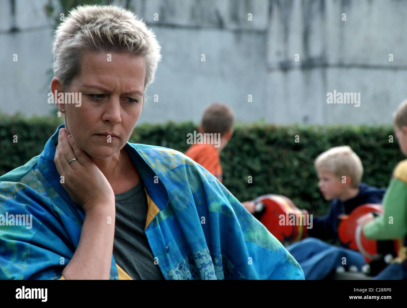 An unhappy adult woman, mum or pedagogical worker sit outside at the children's playground, the worried woman is contemplating, turned her back to the kids. Stock Photo