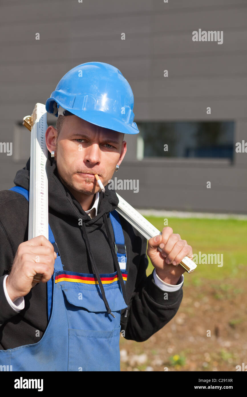 Worker in blue hard hat and workwear holds folding ruler and smokes cigarette Stock Photo