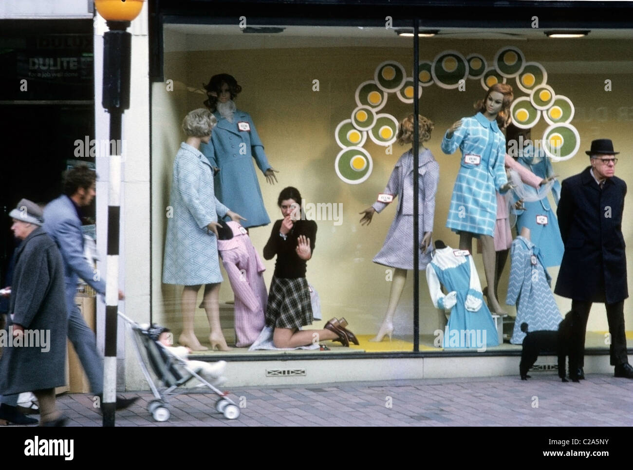 Vintage image of a young woman female shop assistant employee watching people shopping  in the 1970s  working as a window dresser looking out of a women's high street fashion clothing store shop window display, man standing waiting with a black poodle dog and a man walking past in the street with a baby in a McLaren pushchair at Royal Tunbridge Wells town centre in Kent England UK    KATHY DEWITT Stock Photo