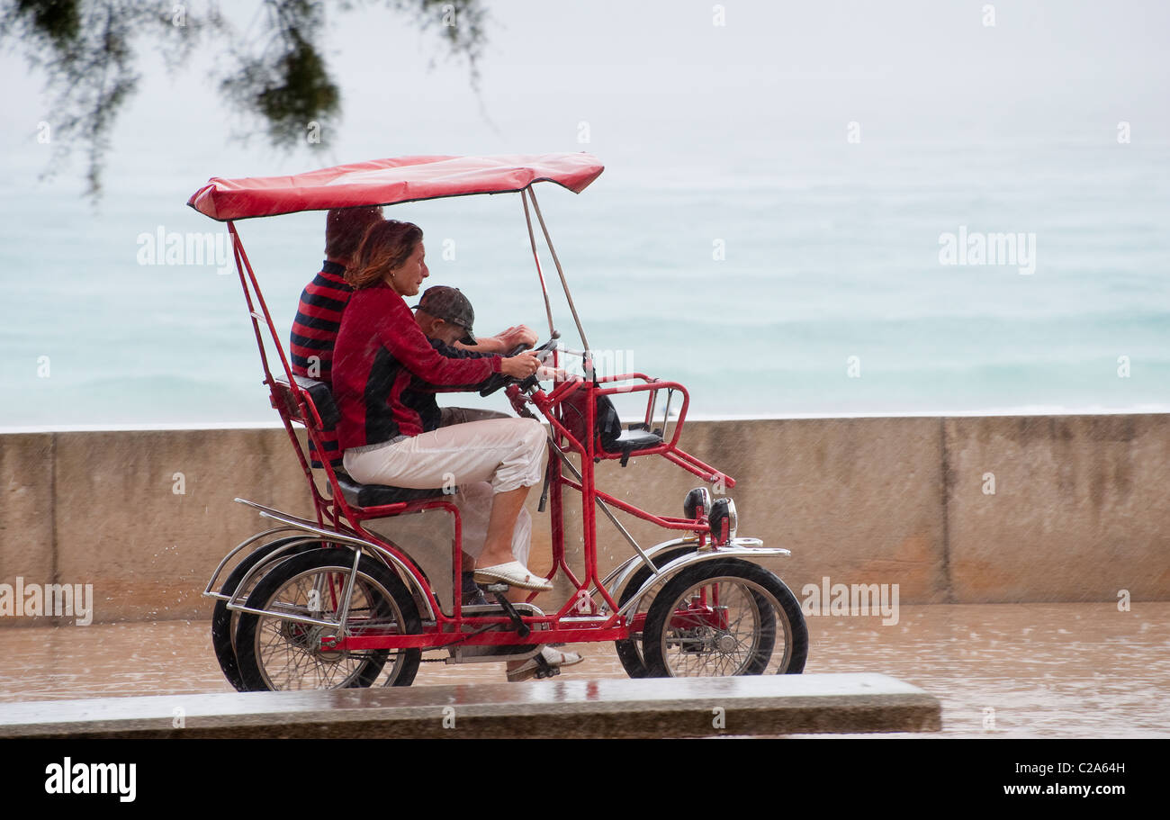 Family using a pedal car bicycle in the rain on holiday in Alcudia,  Mallorca, Spain Stock Photo - Alamy