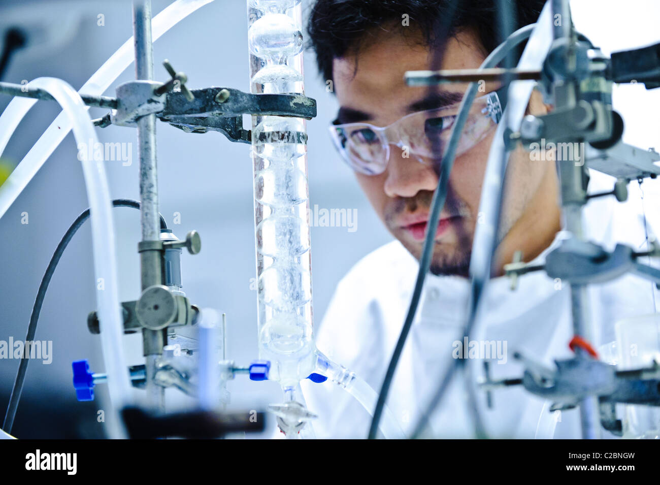 Young Asian male scientist wearing clear goggles and white science coat looking at glass tube apparatus in science lab Stock Photo