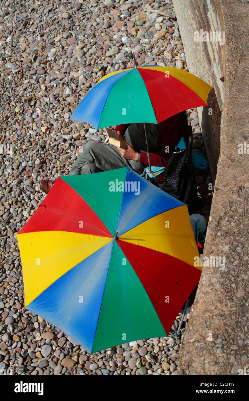 English couple sit on a beach in Devon using colorful sunshades Stock