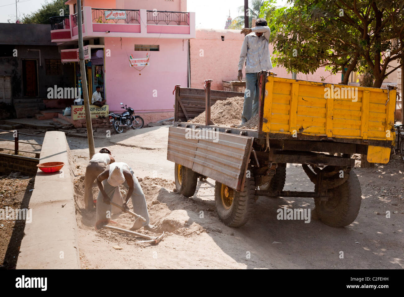 Construction workers shovelling sand beside a yellow tipper truck on a village street, Valsang, Maharashtra, India Stock Photo