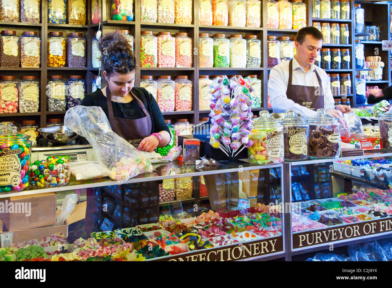 Traditional candy store or sweet shop with display shelving of sweet jars. Two assistants wrapping confectionery at the counter. Stock Photo